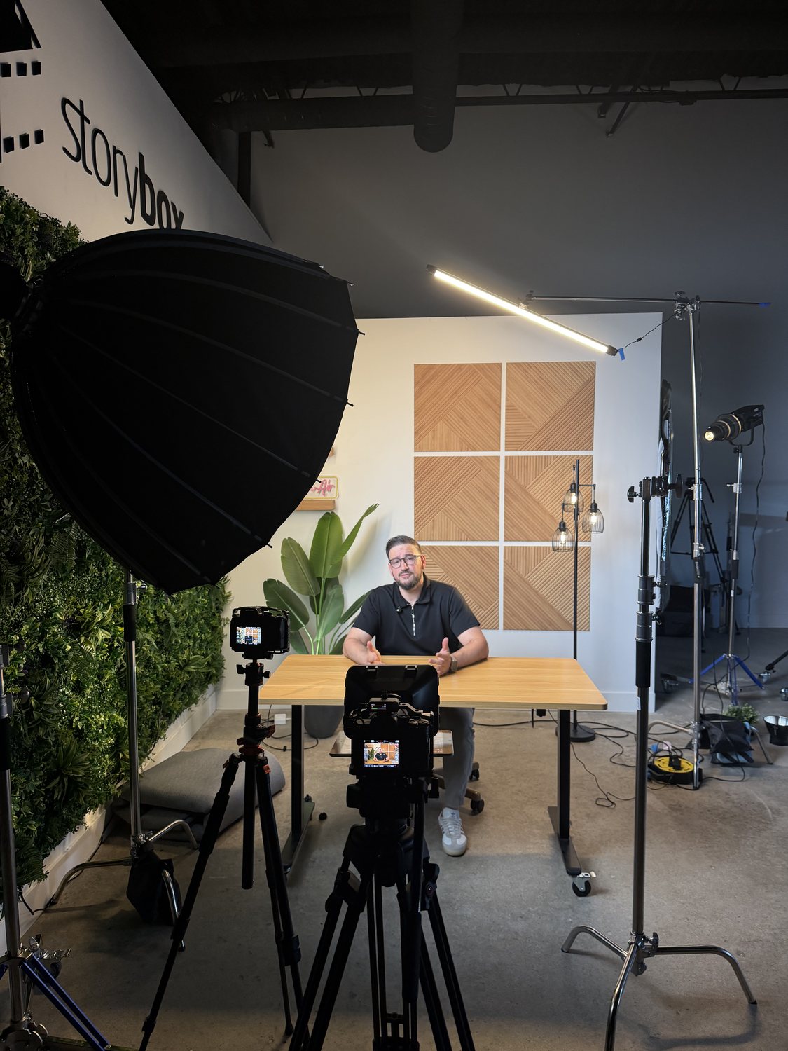A two camera interview setup facing a seated guest at a desk in front of a wood panel backdrop