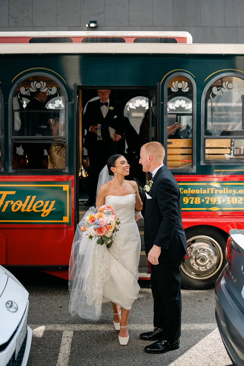 A bride in a strapless wedding dress holds a colorful bouquet while smiling at a groom in a black suit outside a trolley. Guests are seen boarding the trolley behind them.