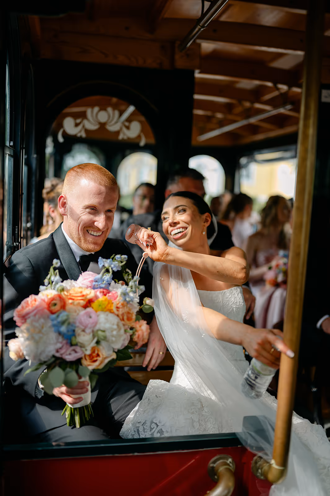 A bride and groom smiling inside a vintage trolley car. The bride is holding a colorful bouquet and playfully touching the groom's face. In the background, guests in wedding attire are seated.