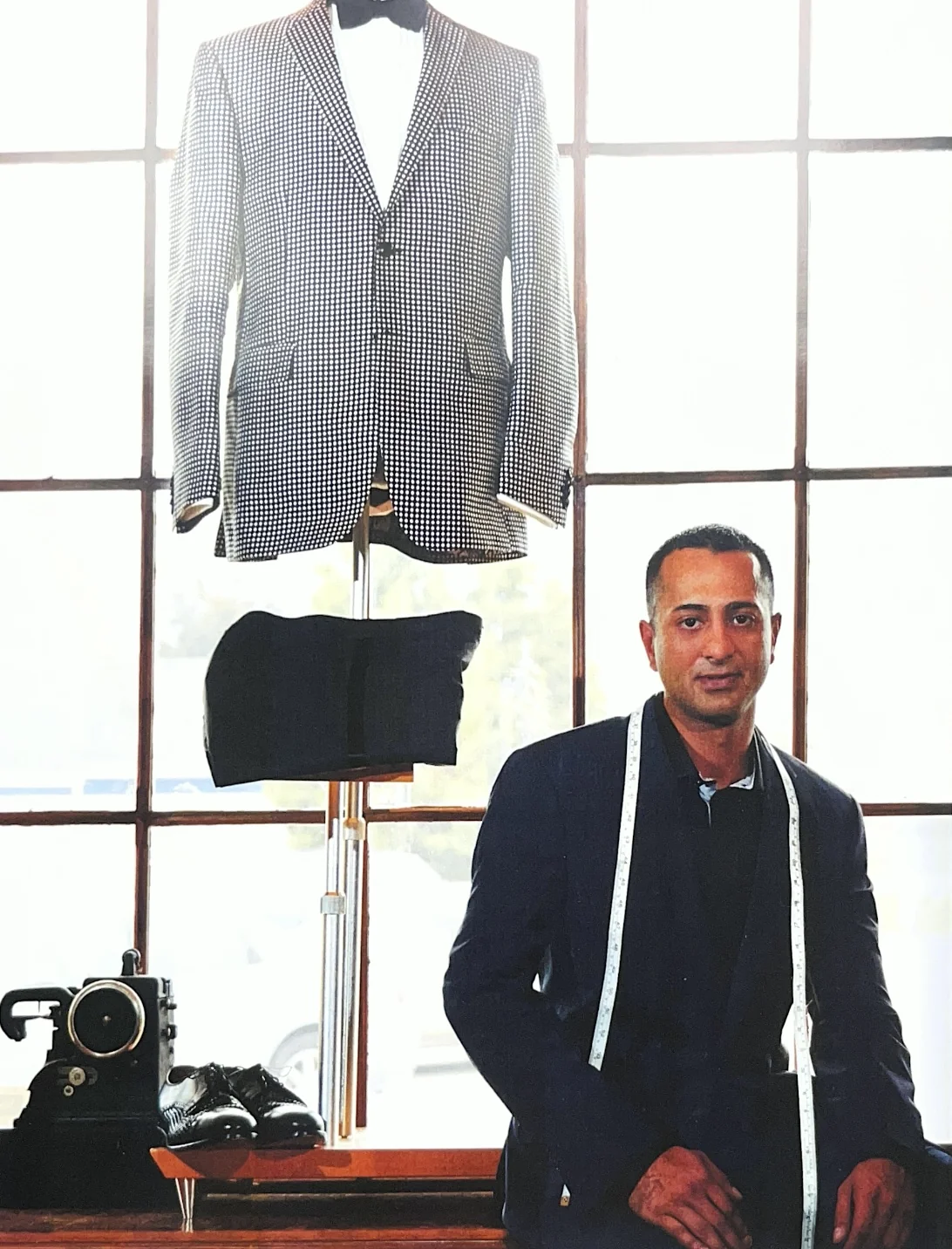 Tailor sitting next to a mannequin dressed in a checkered blazer and black pants, with a sewing machine and shoes on the table beside him.