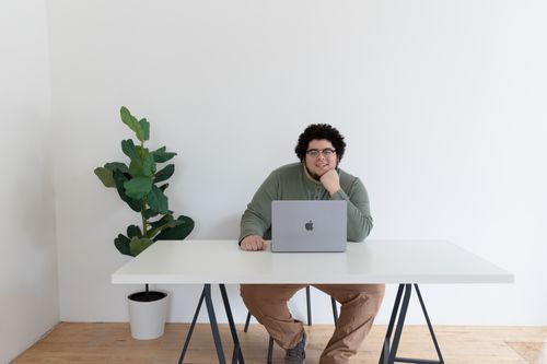 Alex Story portrait brand photo Alex Storey, founder of Skill Mammoth, sitting behind a MacBook at his desk, smiling with a plant in the background, symbolizing friendly and efficient automation expertise.