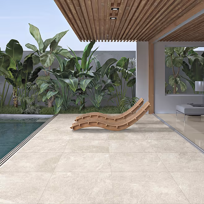 Modern outdoor patio with large beige porcelain tiles, two wooden lounge chairs, a swimming pool, and lush green tropical plants under a wooden slatted roof.