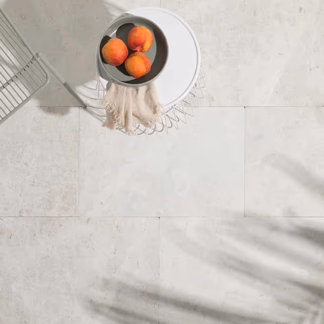 Top view of a white wireframe side table with a gray bowl holding three peaches and a beige cloth, placed on light natural stone tile flooring.