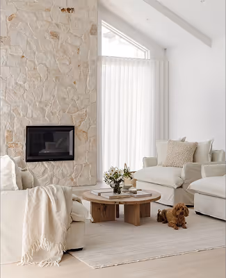 Bright living room with white sofas, a round wooden coffee table, a stone fireplace, and a small brown dog lying on a beige rug.