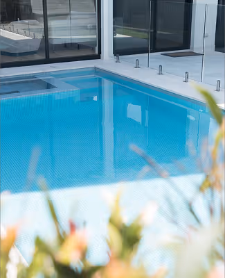 Clear blue outdoor swimming pool next to a modern glass building with blurred plants in the foreground.