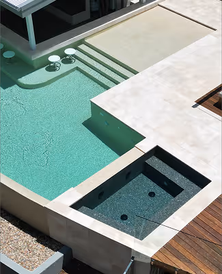 Modern outdoor space with a turquoise swimming pool adjacent to a built-in hot tub surrounded by light stone tiles and wooden decking.