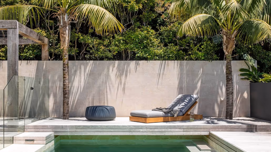 Modern outdoor poolside area with lounge chair, ottoman, palm trees, and glass fence against a beige wall.
