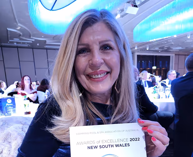 Smiling woman with blonde hair holding an awards certificate for Excellence 2022 from Swimming Pool & Spa Association of Australia, New South Wales.