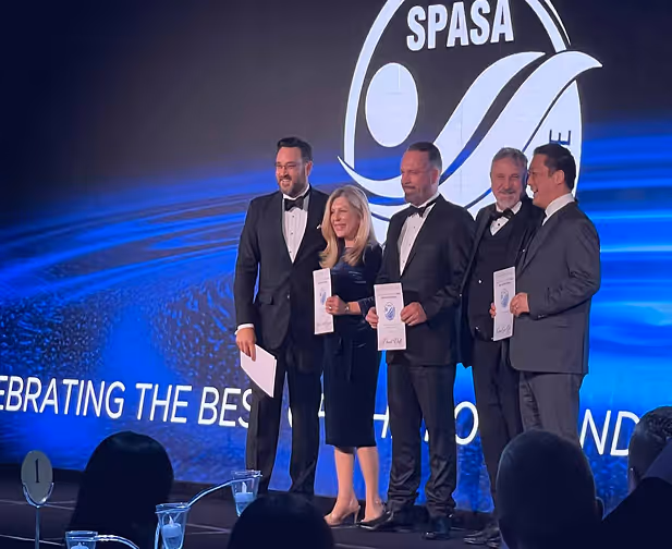 Five people dressed formally in tuxedos and dresses standing on stage holding certificates in front of a SPASA logo and a blue backdrop.