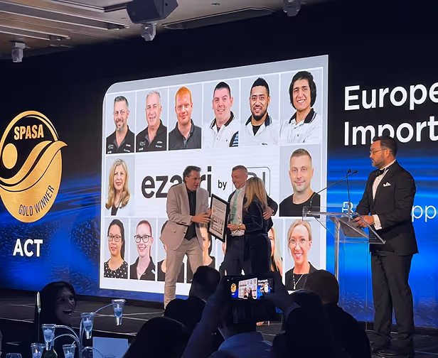 Conference stage with people receiving an award and a large screen showing photos of twelve team members, a gold SPASA ACT winner emblem, and the word 'eza'.