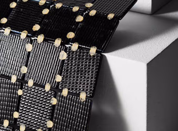 Close-up of a black woven composite material held together by beige rivets or fasteners over a white geometric surface.