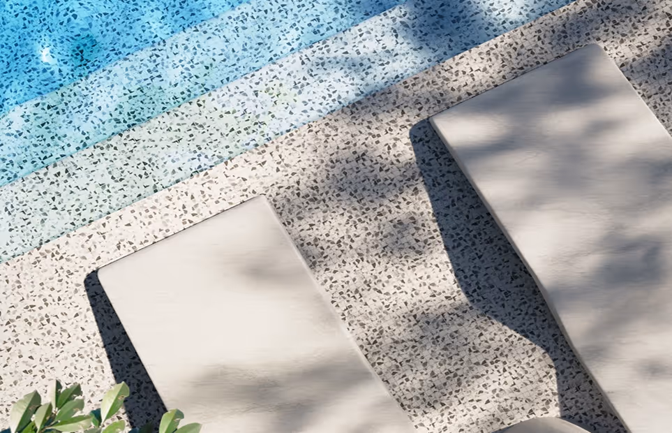 Two beige poolside lounge chairs casting shadows on a tiled pool deck next to clear blue water.