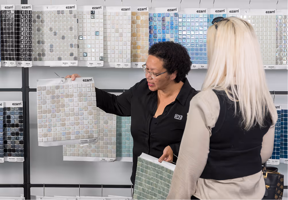 Two women looking at and discussing mosaic tile samples displayed on a wall, one woman holds a tile sheet labeled Ezarri.