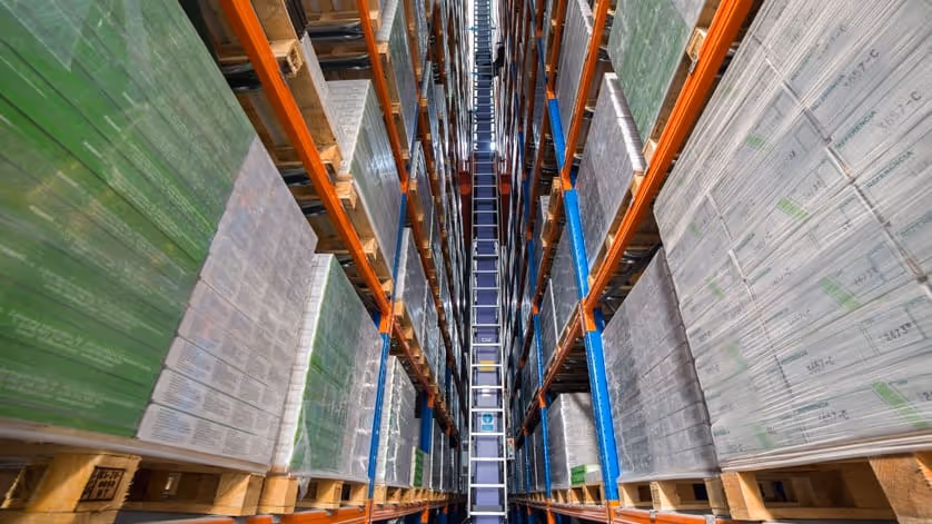 Tall warehouse shelves stacked with pallets of boxes wrapped in plastic, viewed from below looking up between the rows.