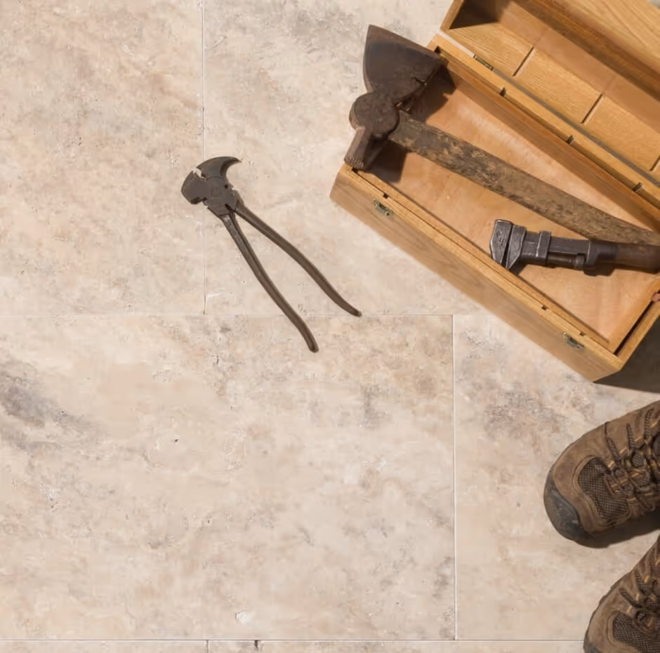 Old hand tools including a hammer, wrench, and pliers on a light stone tile floor next to a wooden toolbox and worn work boots.
