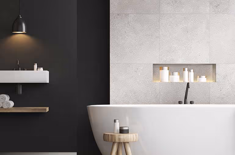 Modern bathroom with a white freestanding bathtub, a wooden stool holding bath products, a wall-mounted sink with toiletries, and a built-in shelf with bottles set in gray tiled wall.