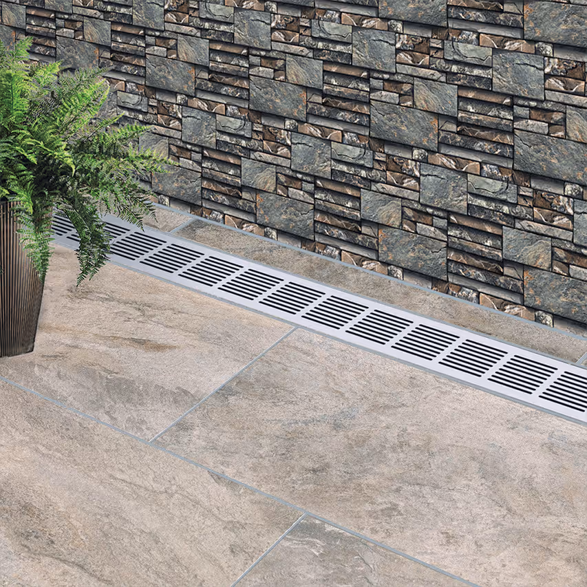 Long stainless steel floor drain with rectangular slots installed along a stone wall with a potted fern nearby on tiled floor.