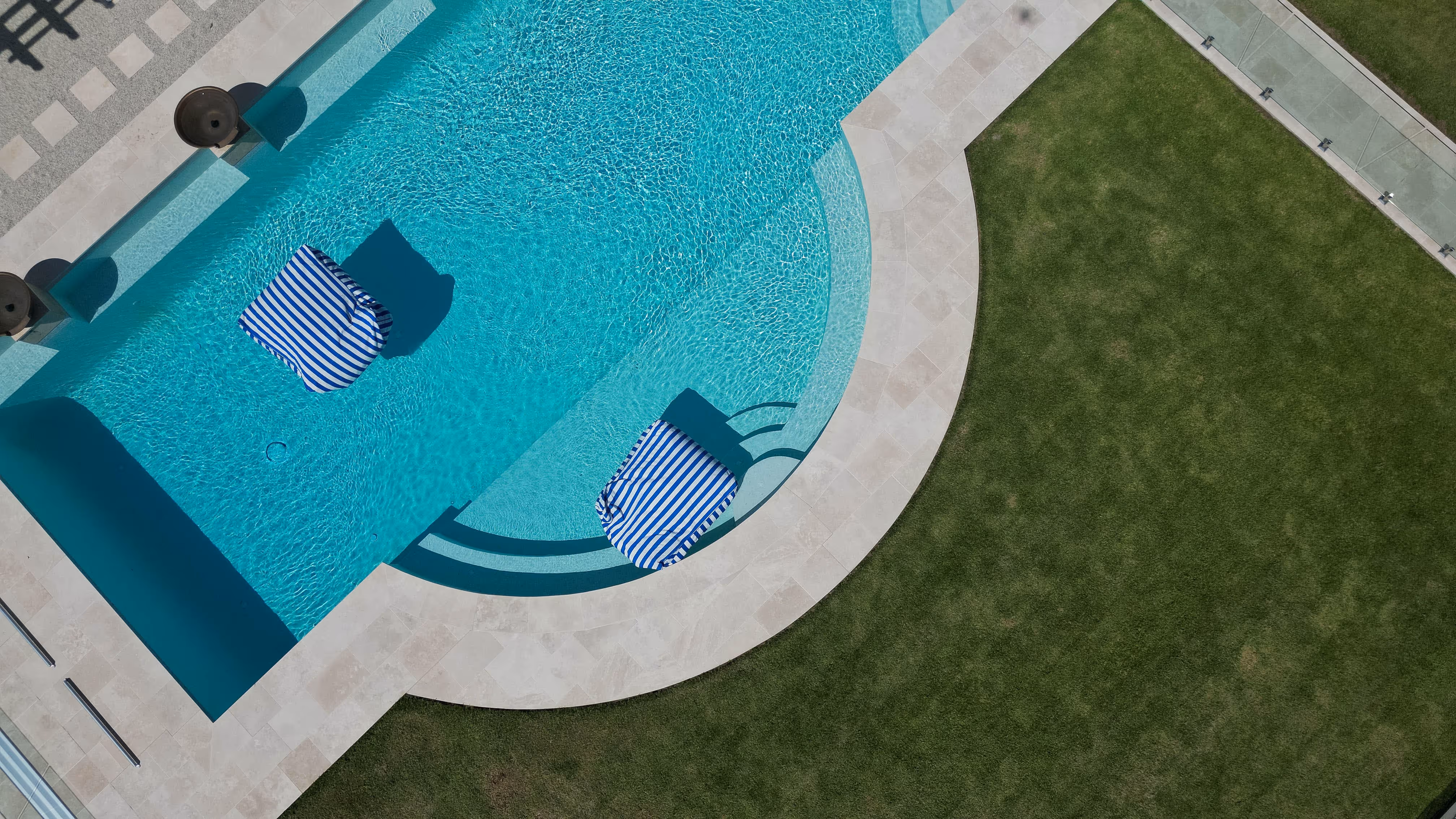 Top-down view of a swimming pool with two blue and white striped lounge chairs partially submerged in clear water next to a grassy lawn.