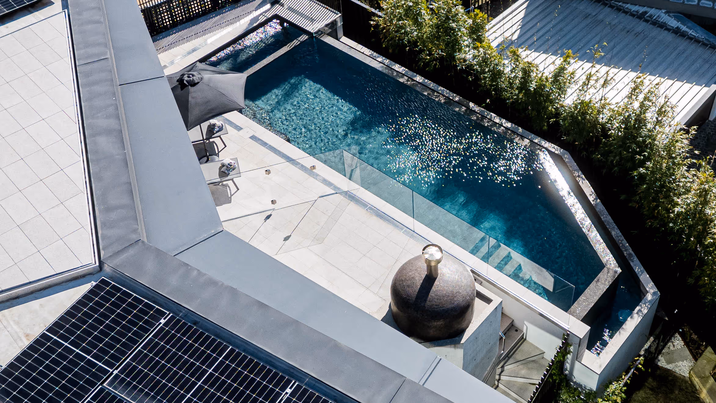 A rooftop view of a modern outdoor pool area with sun loungers, a black umbrella, and solar panels.