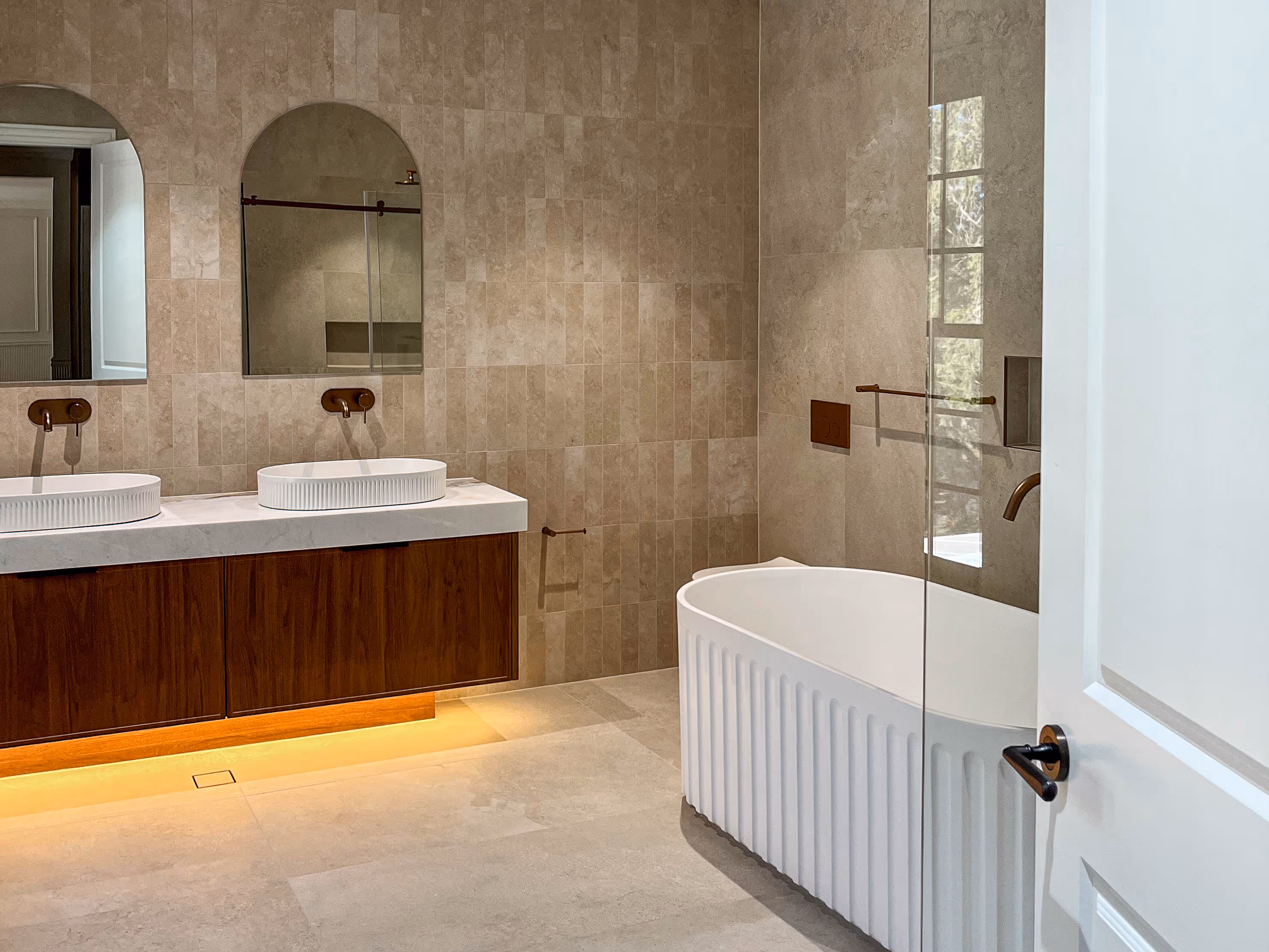 Modern bathroom with freestanding fluted bathtub, double sink vanity with wood cabinet, two arched mirrors, and beige tiled walls and floor.