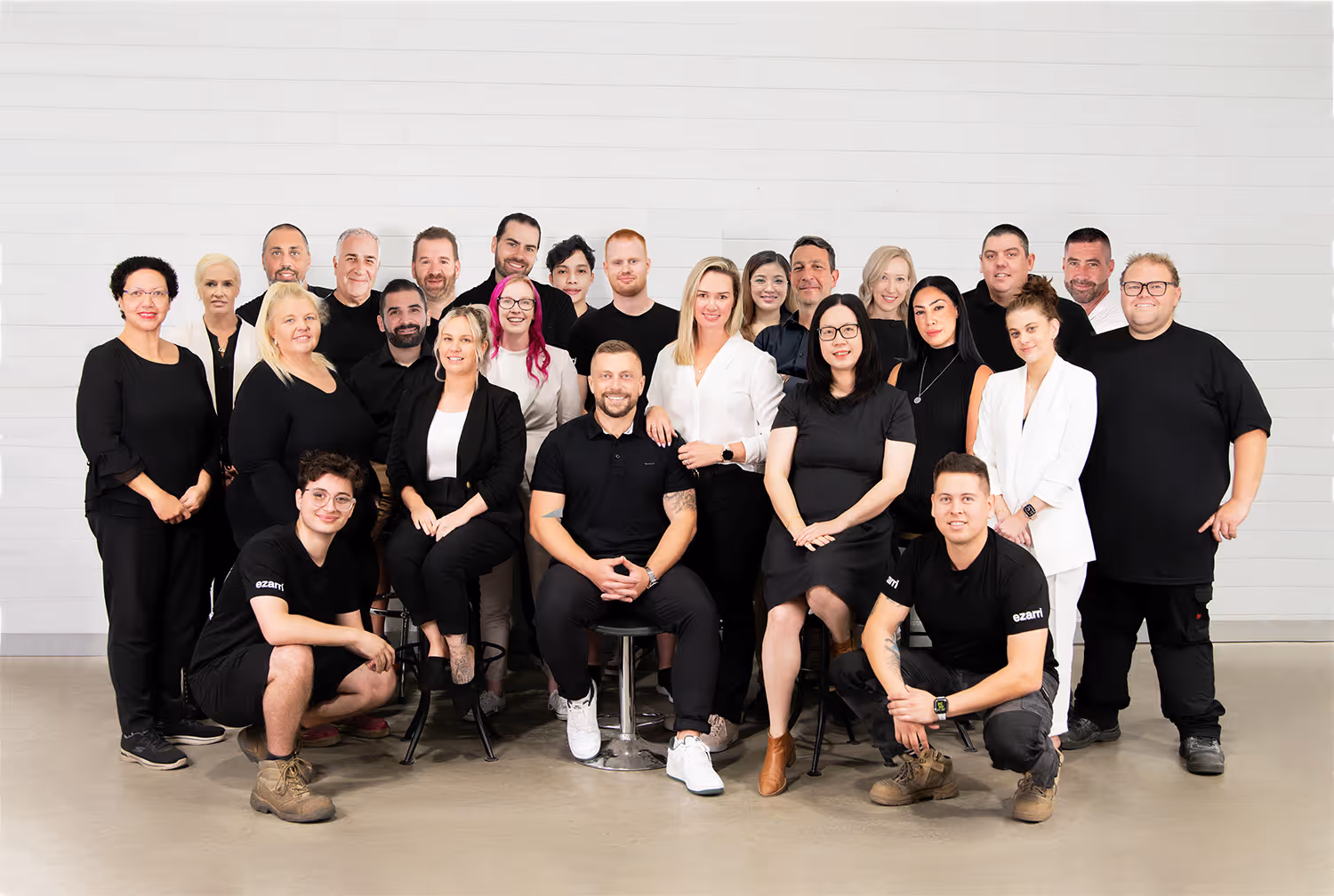 Group photo of 25 diverse team members posing indoors against a white brick wall, wearing mostly black and white clothing.