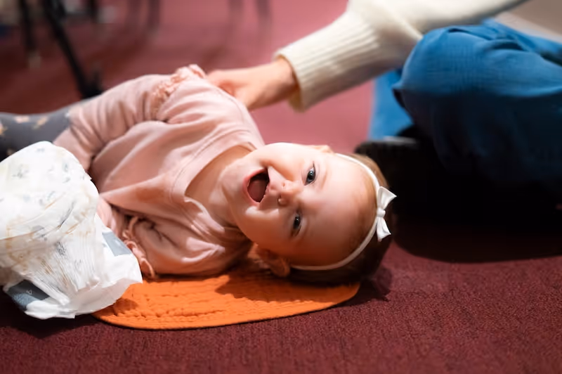 Smiling baby girl lying on her side on an orange mat, wearing a pink outfit and white headband with a bow.