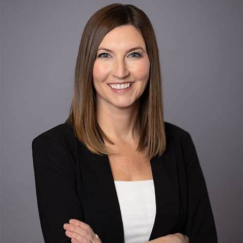 Portrait of Lauren Polovoy, Certified Nutrition Coach and Group Fitness Trainer, smiling and wearing a black blazer over a white top against a gray background.
