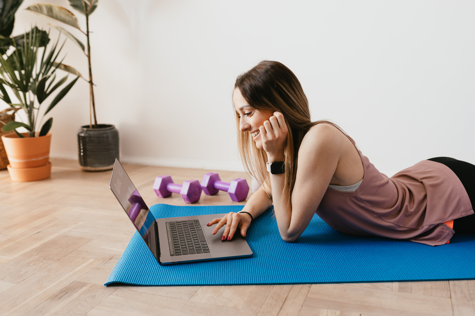 Woman on a yoga mat using a laptop for a wellness coaching session, with dumbbells nearby, representing assessment of health goals."