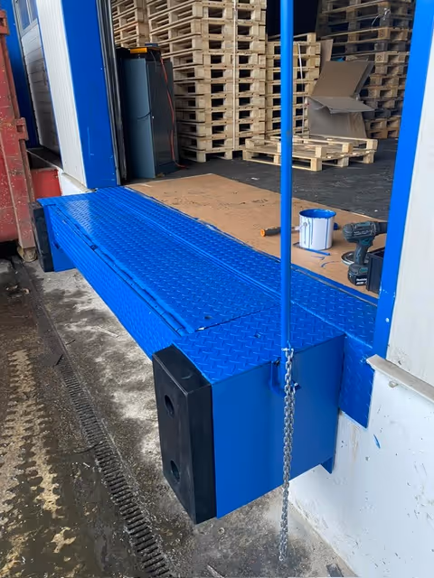 Blauer Verladerampe mit Gummipuffer vor Lagerraum mit gestapelten Holzpaletten und Malerbedarf.