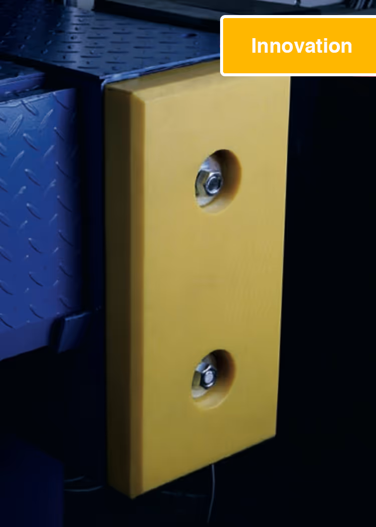 Nahaufnahme eines gelben, rechteckigen Schutzpuffers mit zwei sichtbaren Metallbolzen an einer schwarzen Metallstruktur.