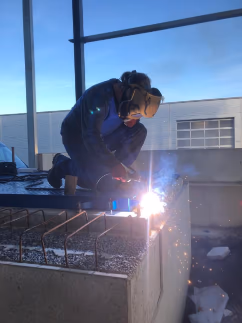 Person trägt Schutzhelm und schweißt an einer Betonstruktur mit sichtbaren Funken und Rauch.