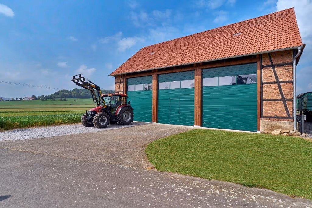 Landwirtschaftlicher Traktor steht neben einem großen Gebäude mit rotem Ziegeldach und grünen Toren auf gepflastertem Hof.