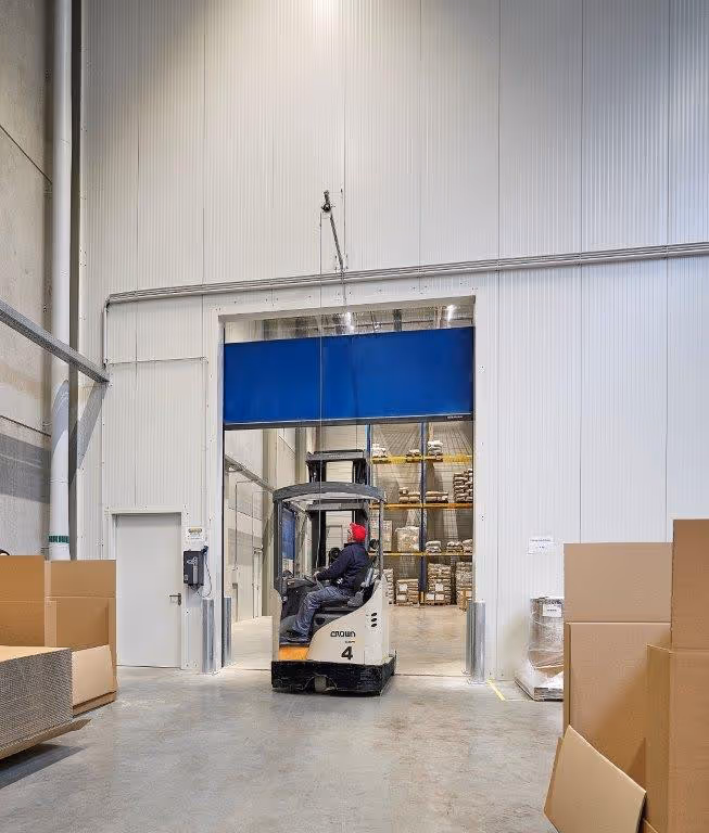 Gabelstapler fährt durch ein hohes, blaues Schnelllauftor in einem Lager mit Kartons und Regalen voller verpackter Waren.