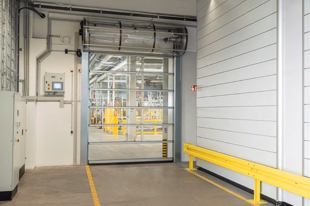 Transparente industrierolltor im geöffneten Zustand in einer Lagerhalle mit gelber Schutzbarriere und Steuerungskasten an der Wand.