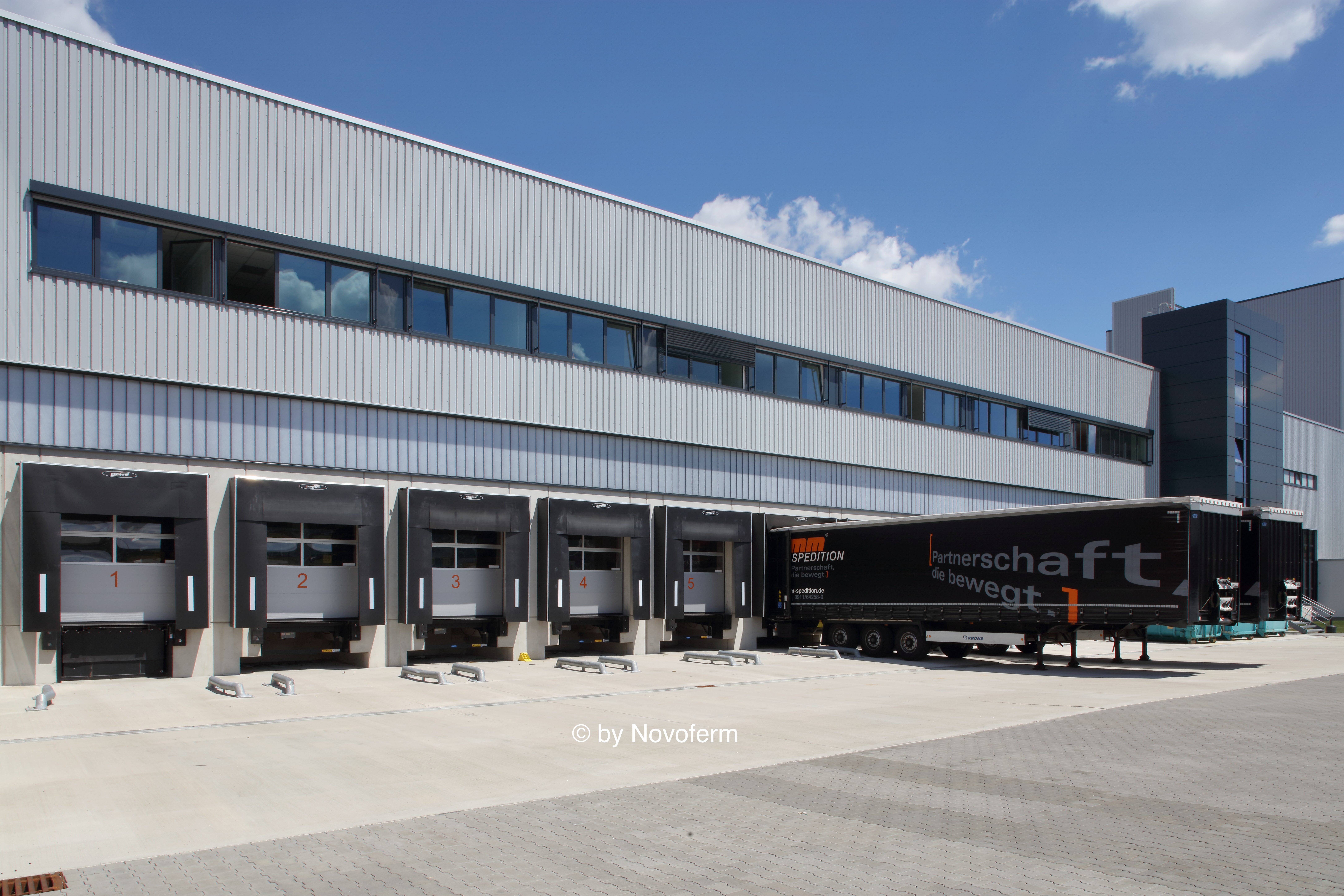 Sechslädtige Lagerhalle mit anhängendem schwarzen LKW-Auflieger unter klarem blauem Himmel.