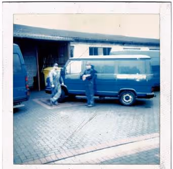 Zwei Personen neben einem blauen Transporter vor einem Garagentor auf einem gepflasterten Hof.