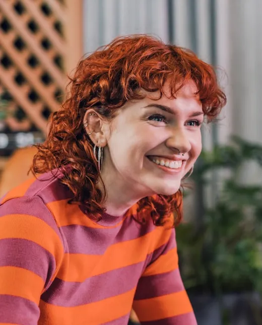 Junge Frau mit roten lockigen Haaren und septum Piercing lächelt, trägt gestreiften Pullover in Orange und Lila.