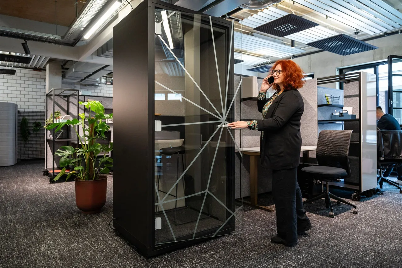 Frau mit roten Haaren telefoniert in einer modernen Büroeinheit mit Glaswand und offenem Arbeitsbereich.