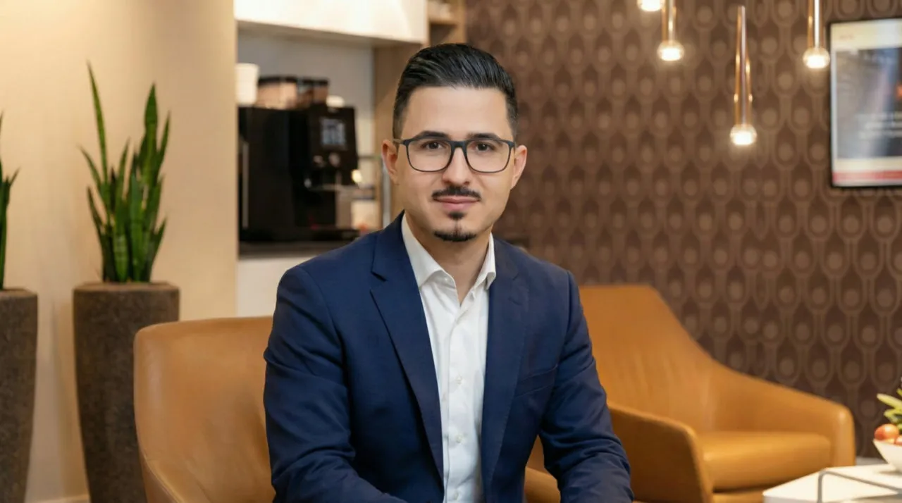 Mann mit dunklen Haaren, Brille und blauem Anzug sitzt in einem Lederstuhl in einem modernen Lounge-Bereich.