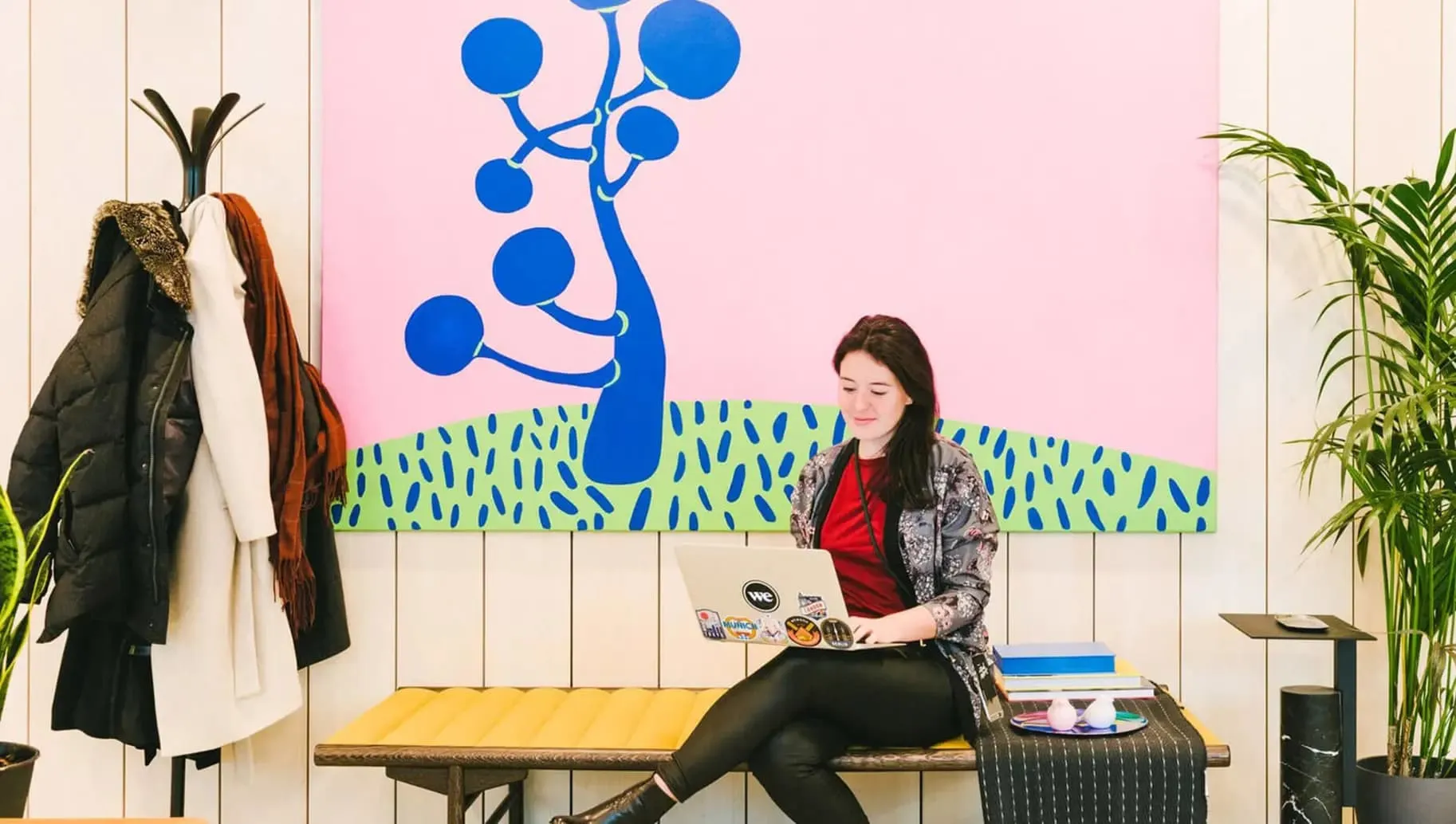 Eine Frau sitzt auf einer Bank mit einem Laptop vor einer bunten Wand mit abstraktem Baum, umgeben von Pflanzen und einer Garderobe mit Mänteln.