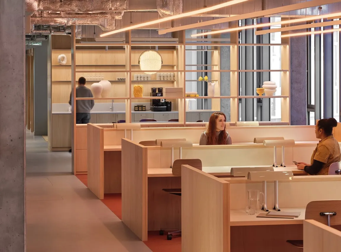 Modern office with wooden walls, built-in workplaces and two women talking, in the background a man at the coffee station.