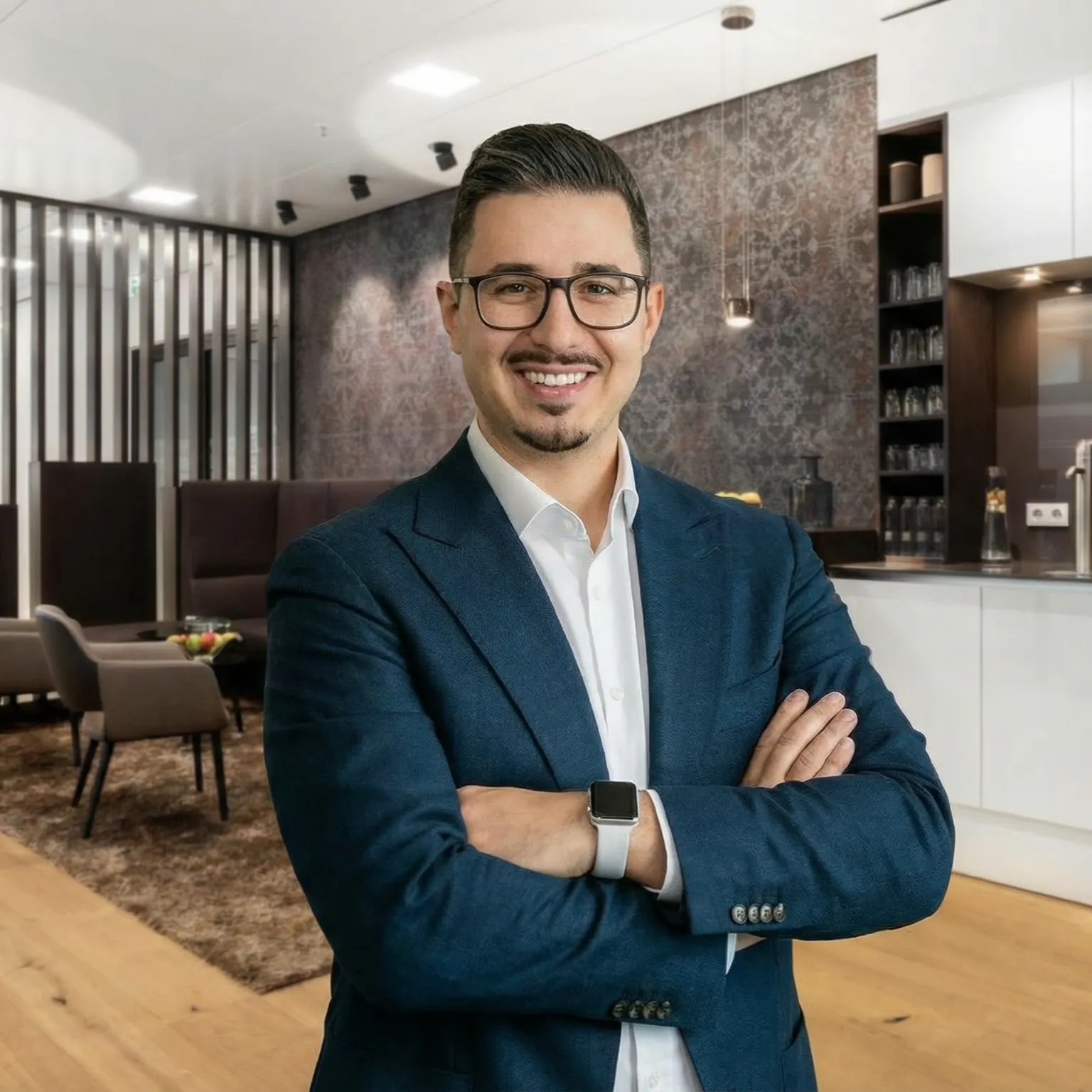 Lächelnder Mann mit Brille und blauem Anzug in modernem Büro mit überkreuzten Armen.