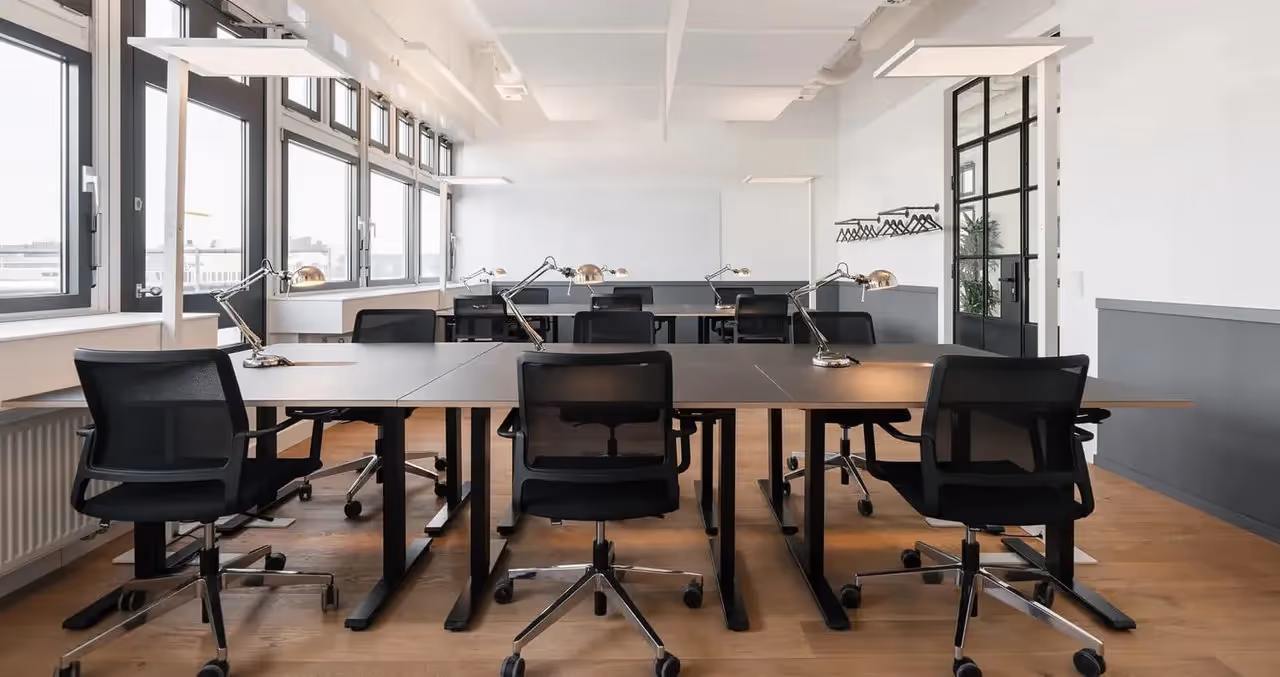 Mehrpersonenbüro entlang großer Fenster, graue Tische, schwarze Drehstühle, verchromte Lampen