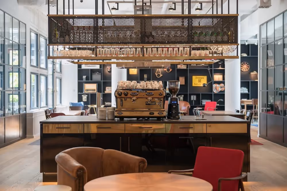 Kaffeebar mit goldfarbener Espressomaschine, hängenden Gläserregalen, dunklen Fronten, braunen und roten Sesseln