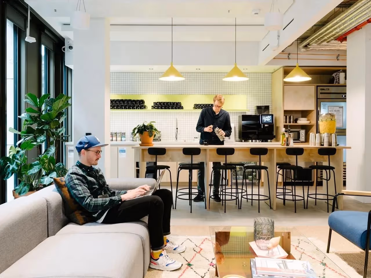 Moderne Lounge mit Sofa, Pflanzen, Barhockern, Theke, zwei Personen; helle Pendelleuchten, offene Regale.