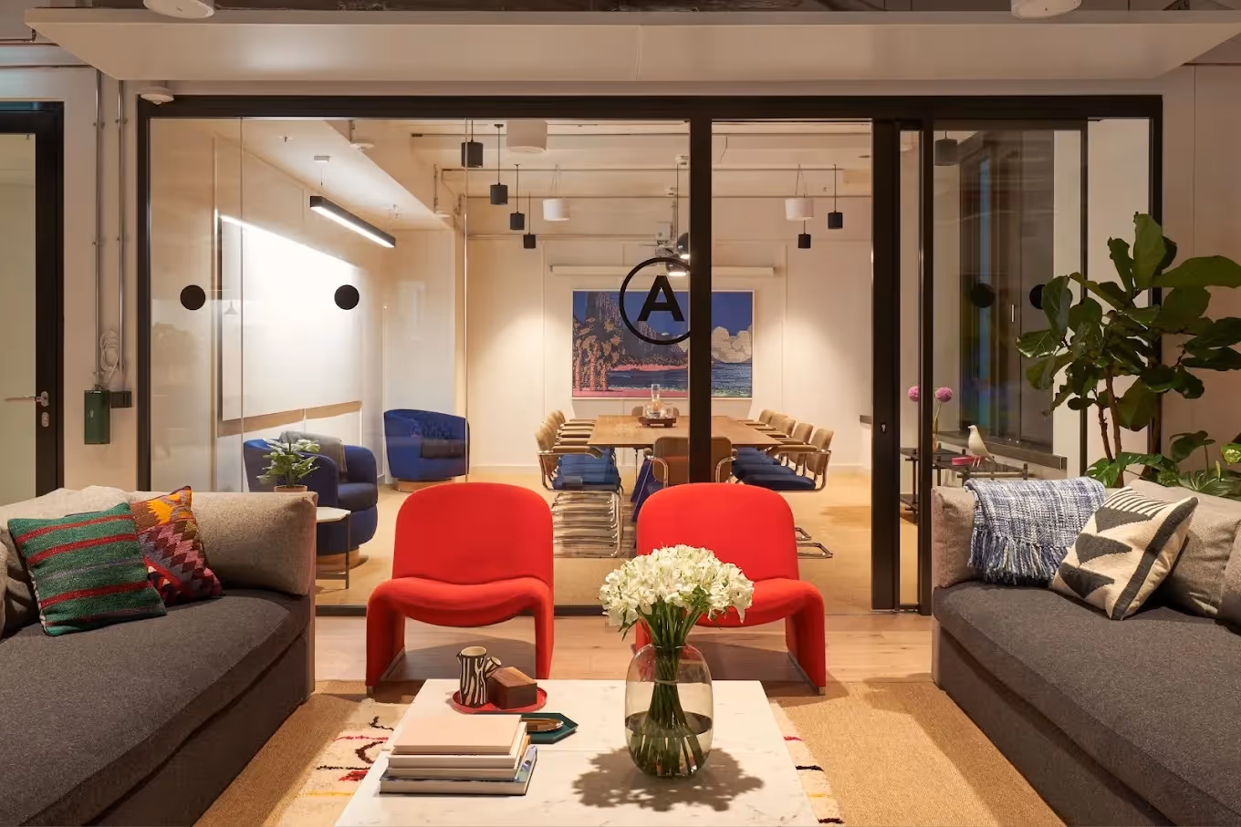 Graue Sofas, rote Stühle, Glastisch mit Blumen, Konferenzraum mit Holztisch und blauen Sesseln im Hintergrund.