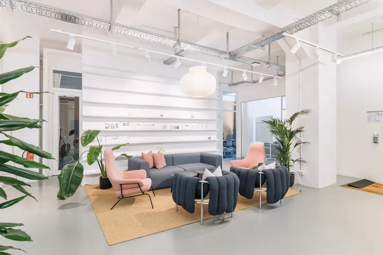 Heller Raum mit grauem Sofa, rosa Sesseln, Pflanzen, Regalen und Hängelampe auf Teppichboden.