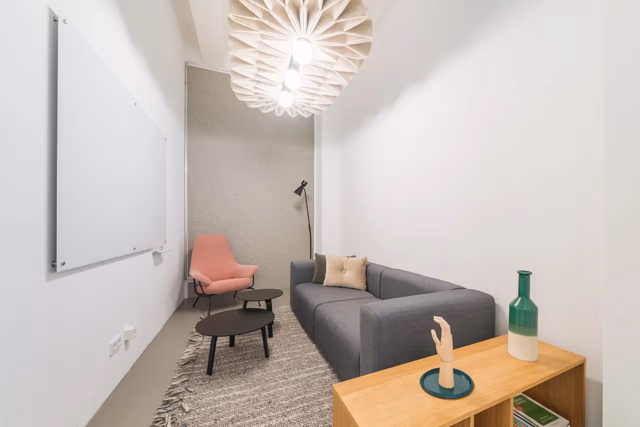 Graues Sofa, rosa Sessel, weiße Wände, große Lampe, Holztisch mit Deko, Teppich, Whiteboard an der Wand.