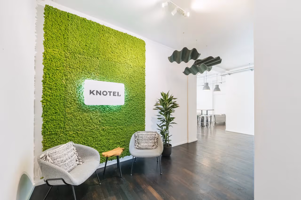 Grüne Mooswand mit Knotel-Logo, zwei graue Sessel, Holzbeistelltisch, große Pflanze, dunkler Holzboden, moderne Lampen.