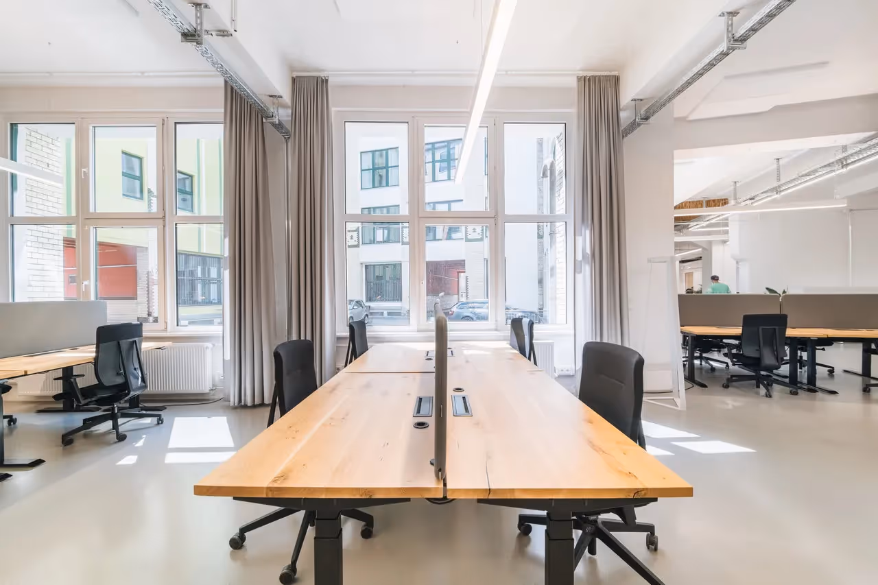 Helles Büro mit großen Fenstern, Holztischen, schwarzen Bürostühlen und grauen Vorhängen, offene Raumgestaltung.